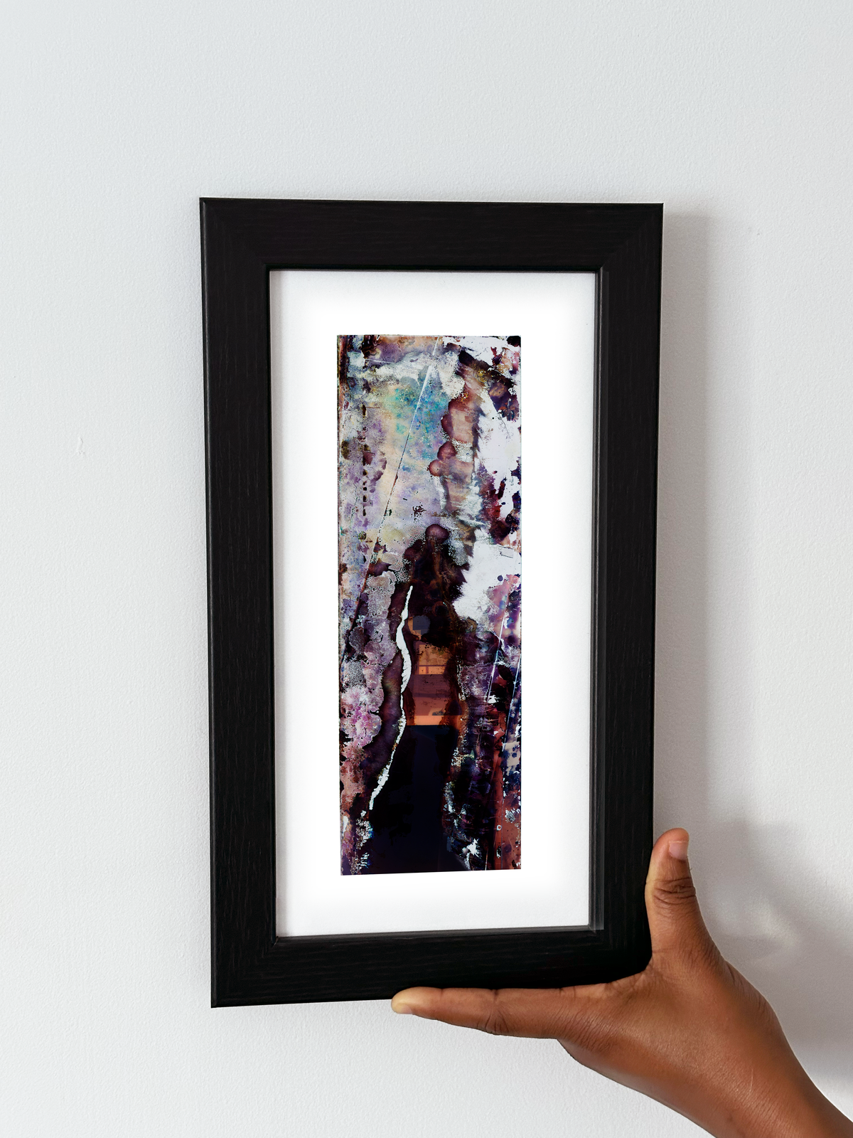 A hand holding a black-framed abstract art print with a white mat, featuring Naturalia II #029 against a plain white wall.