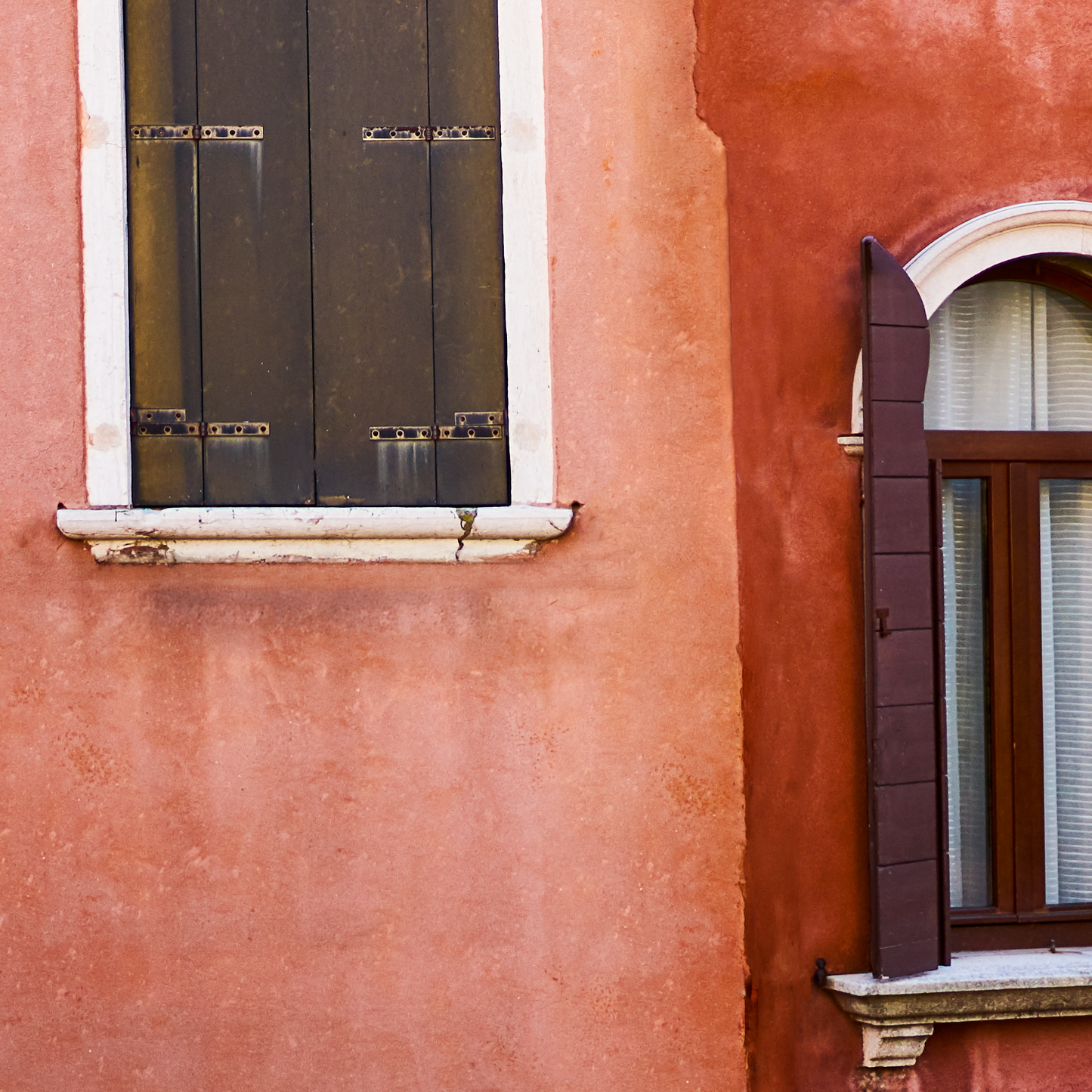 Close-up of a red building with two windows, one with black shutters and the other with brown shutters.