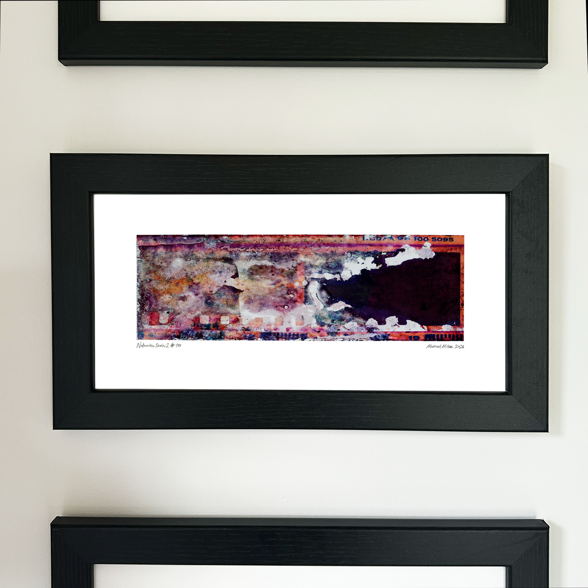 A framed abstract art print with a colorful textured design in red, purple, and white, set within a black frame and mounted on a light beige wall.
