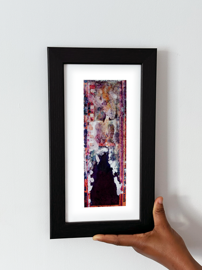 A hand holding a black-framed abstract art print with a white mat against a plain white wall.