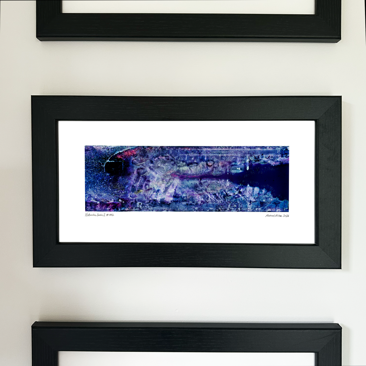 A framed panoramic art print with a black frame and white mat, featuring an abstract blue, purple, and black aerial-style landscape design, mounted on a white wall.