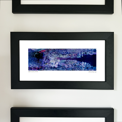 A framed panoramic art print with a black frame and white mat, featuring an abstract blue, purple, and black aerial-style landscape design, mounted on a white wall.