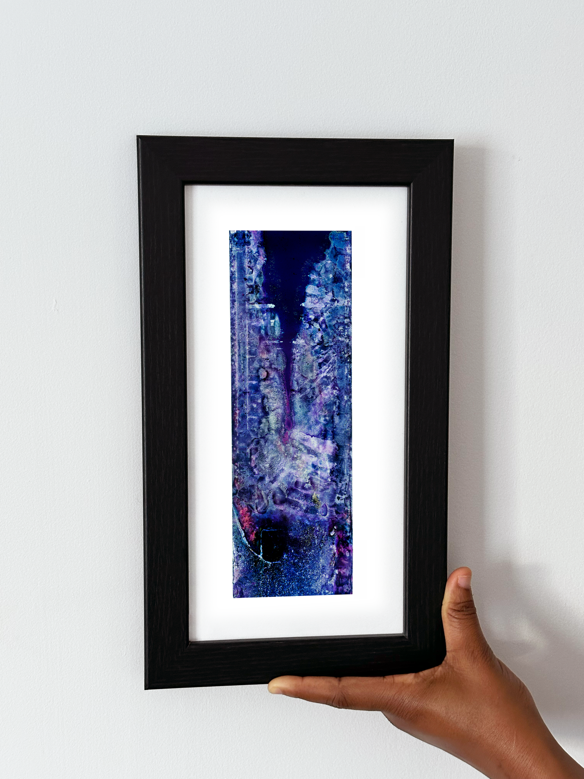 A hand holding a black-framed abstract art print with a white mat. The artwork features a vertical composition with deep blue, purple, and white textured patterns.