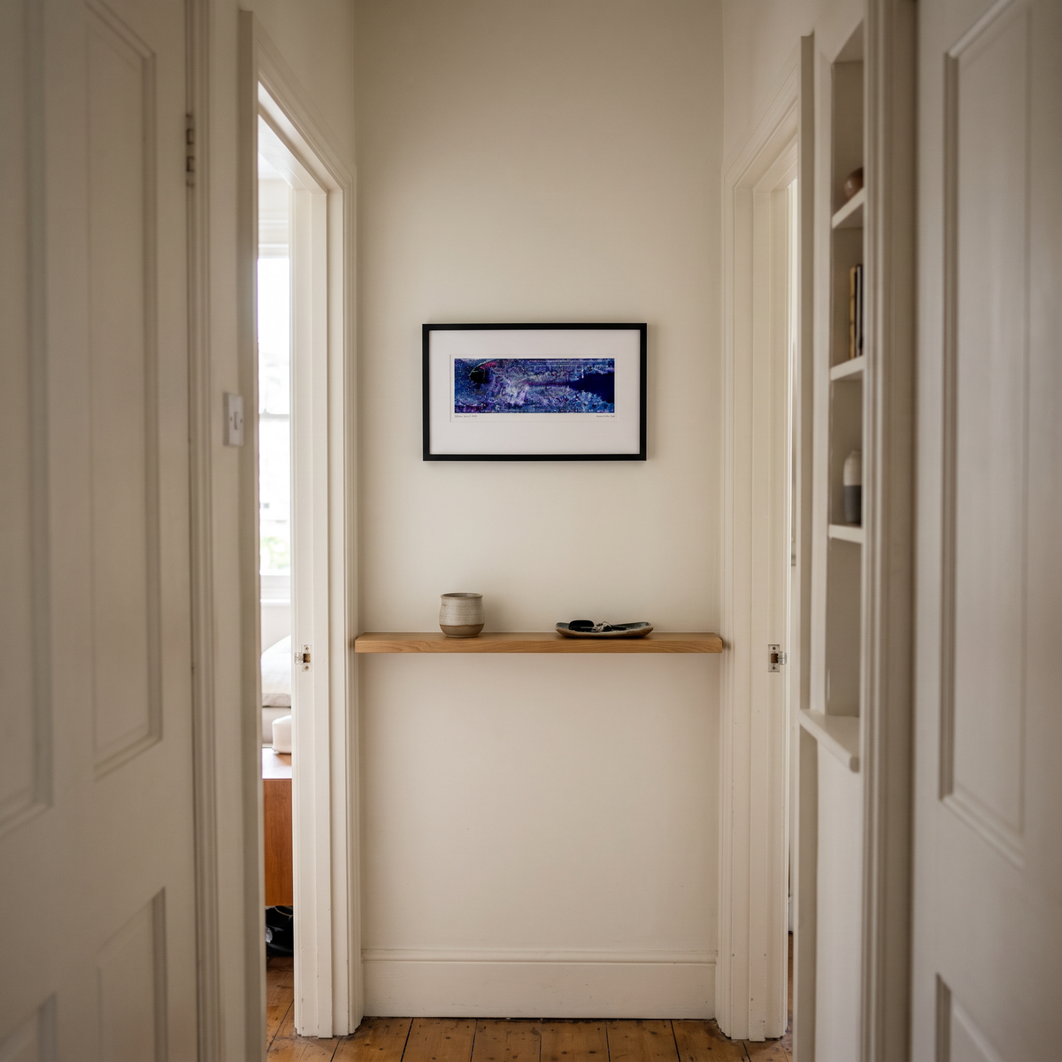 A framed horizontal landscape art print with blue and purple tones hangs on a cream-colored wall above a light wood console shelf in a home hallway.