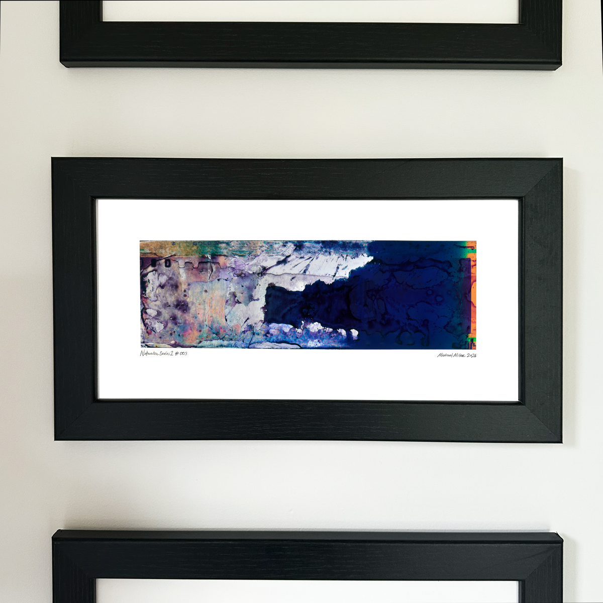 A framed abstract art print with a black frame and white mat, displayed on a light wall. The artwork features a dark blue textured area on the right with splashes of purple, pink, and green on the left.