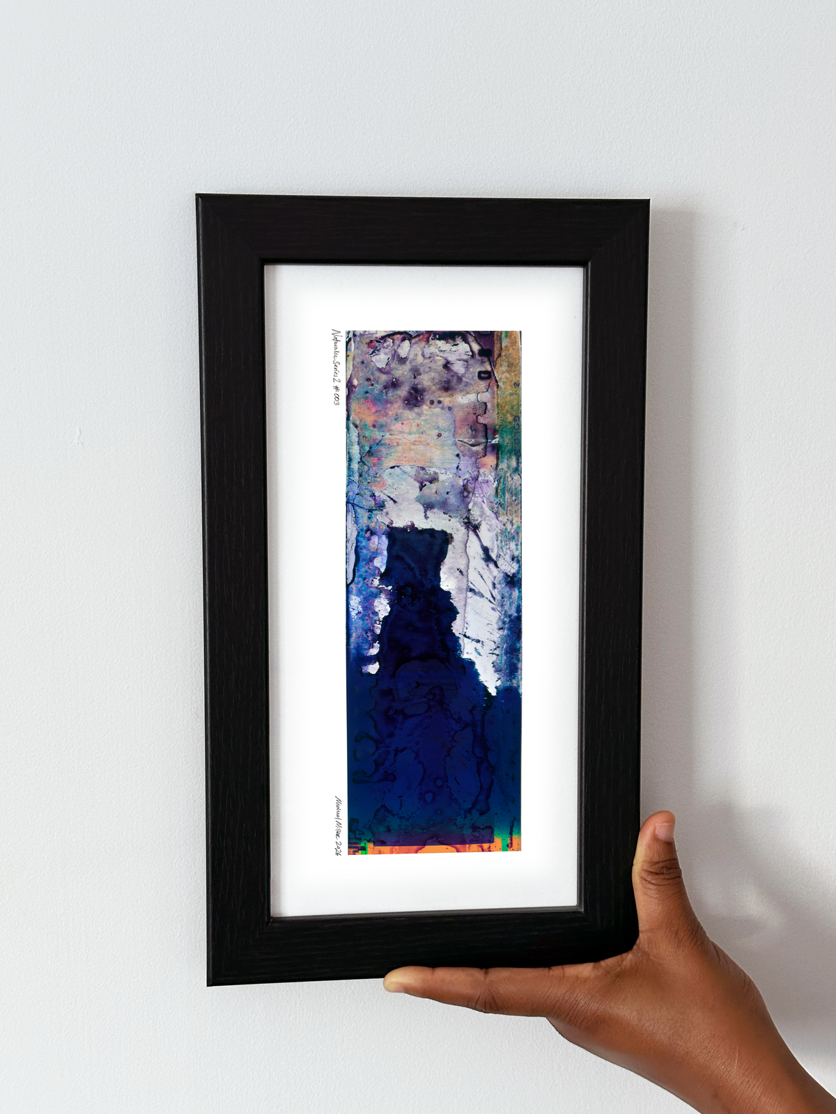 A hand holding a black-framed abstract art print with a white mat, set against a plain white wall.