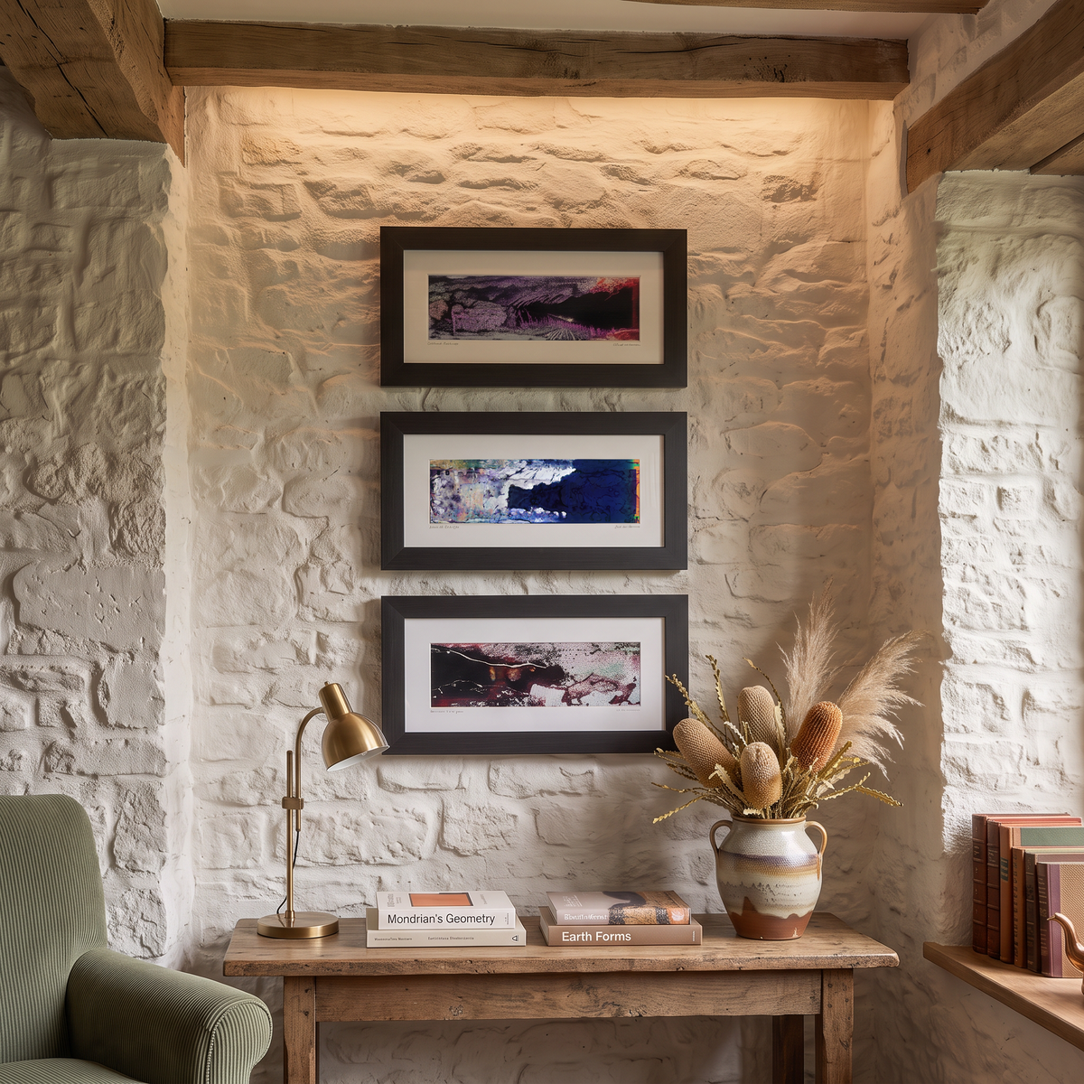 Three framed landscape art prints with black frames are displayed vertically on a white stone wall above a rustic wooden console table in a cozy room setting.