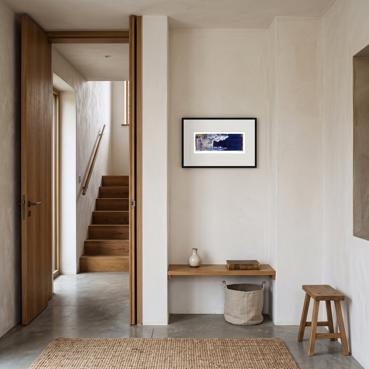 A framed horizontal art print hangs on a cream-colored wall in a minimalist, modern interior hallway with wooden stairs, a bench, and a stool.