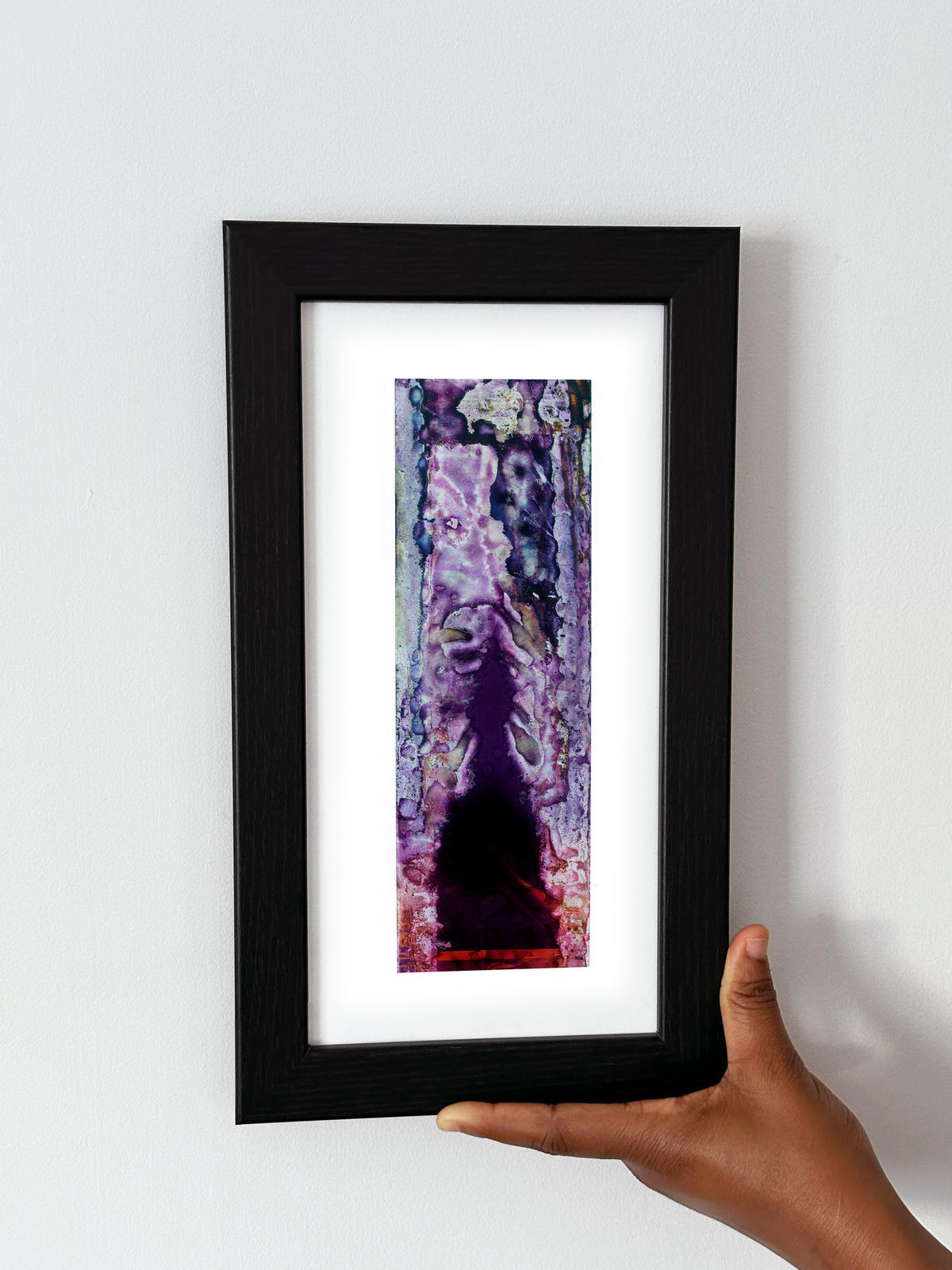 A framed abstract art print featuring purple, black, and pink tones, held by a hand against a plain white wall.