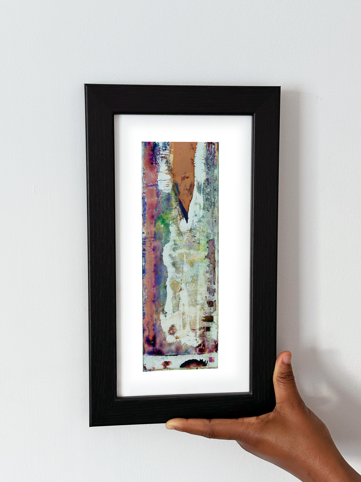 A hand holding a black-framed abstract art print with a white mat, featuring Naturalia II #005 against a plain white wall.
