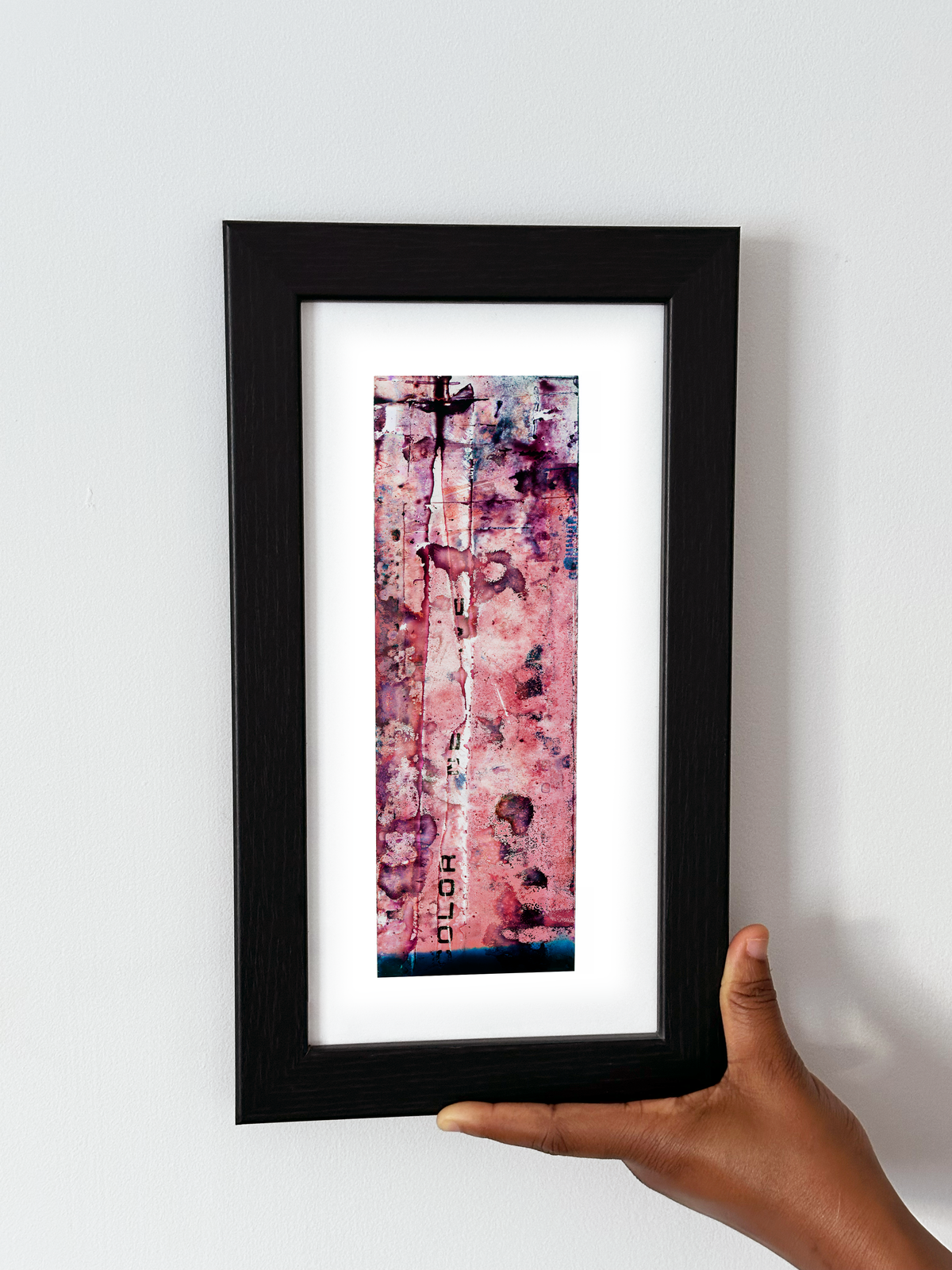 A hand holding a black-framed abstract art print with a white mat, featuring Naturalia II #006 against a plain white wall.
