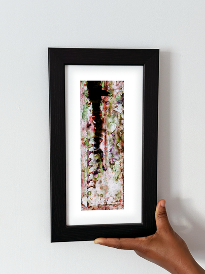 A hand holding a black-framed abstract art print with a white mat, featuring Naturalia II #007 against a plain white wall.
