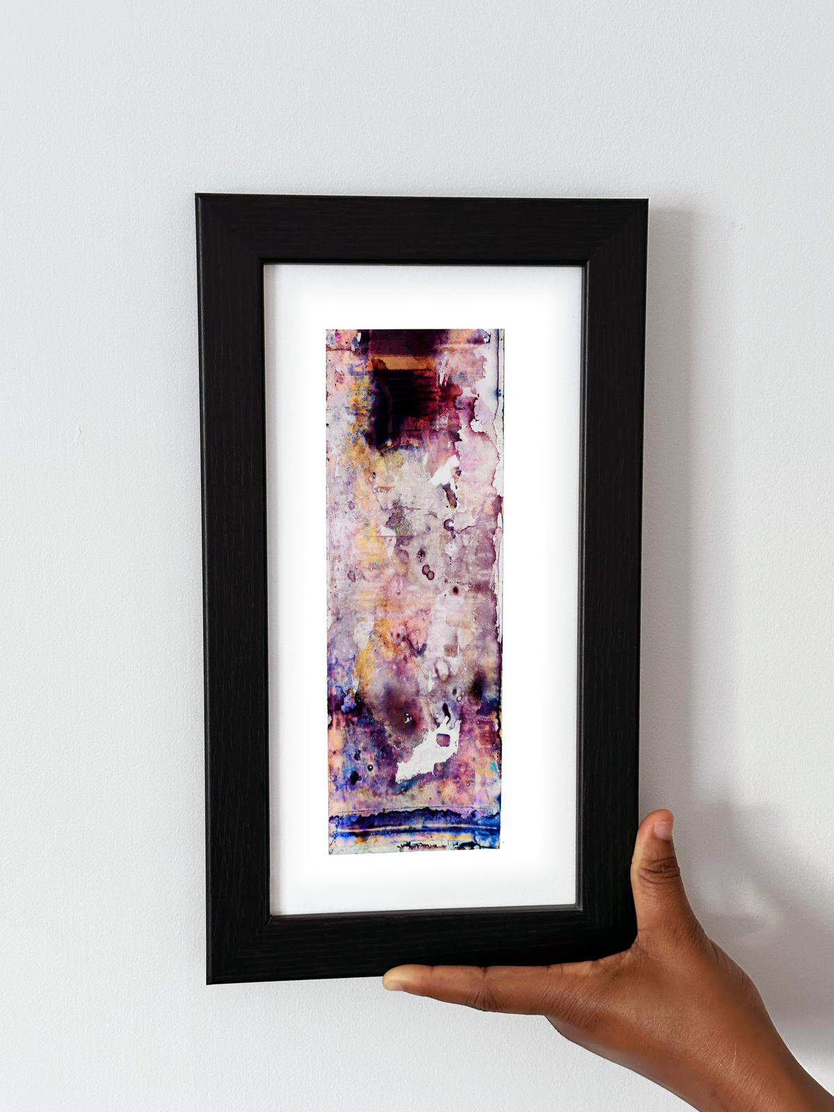 A hand holding a black-framed abstract art print with a white mat, featuring Naturalia II #008 against a plain white wall.

