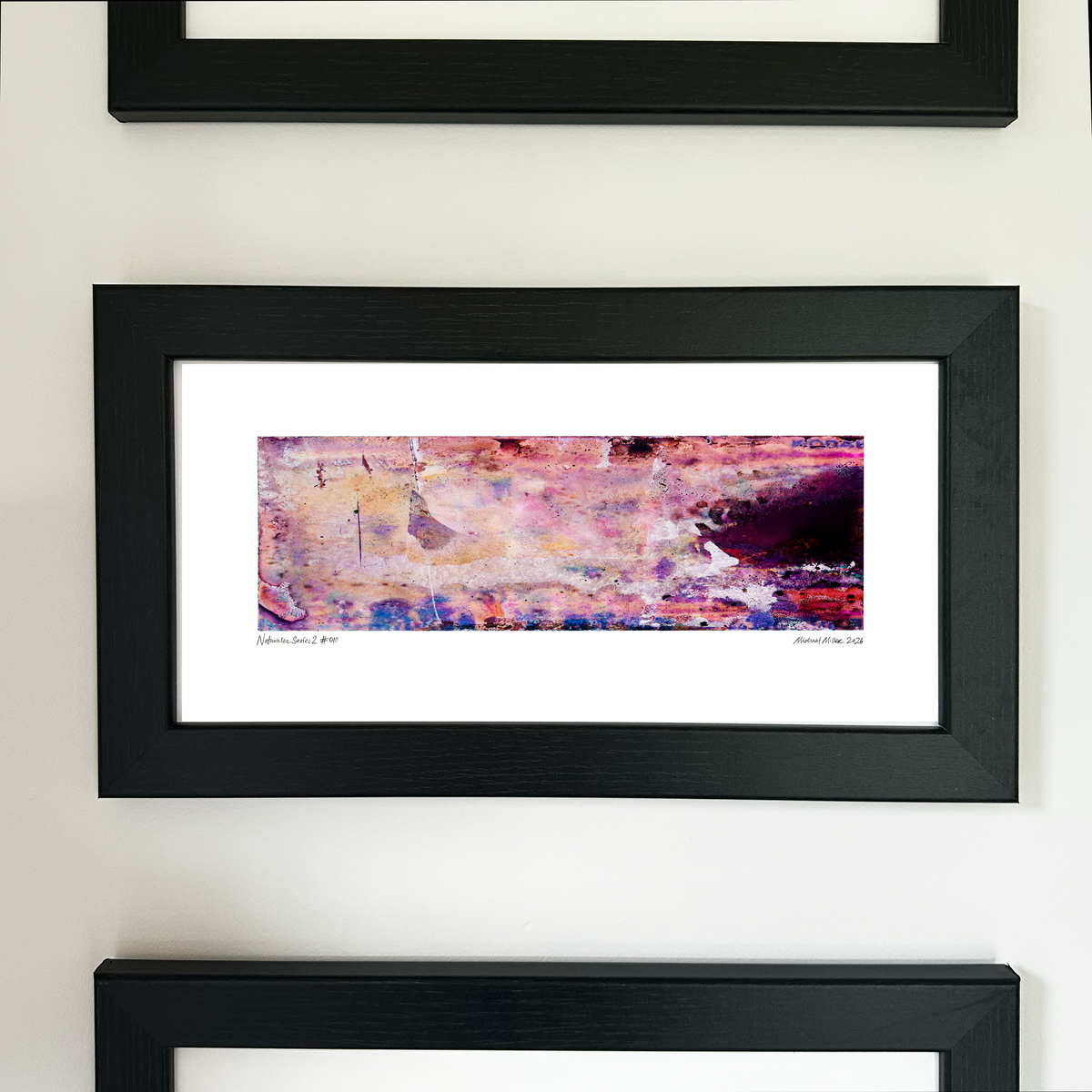 A framed abstract art print with a black frame and white mat, featuring Naturalia II #010 in pink, peach and purple tones, mounted on a white wall.

