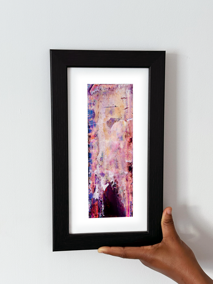 A hand holding a black-framed abstract art print with a white mat, featuring Naturalia II #010 against a plain white wall.
