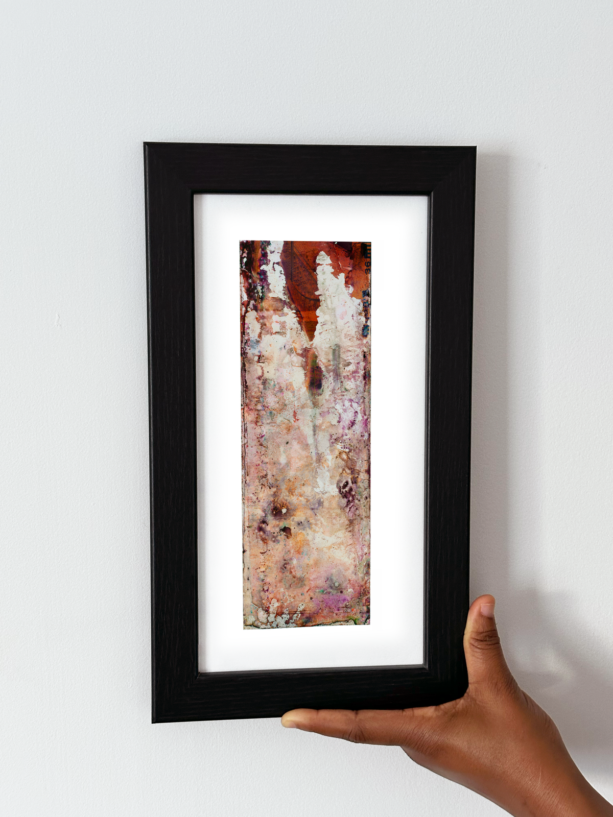 A hand holding a black-framed abstract art print with a white mat, featuring Naturalia II #012 against a plain white wall.
