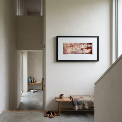 A framed panoramic abstract art print in pink, peach, orange and purple tones, displayed on a wall in a styled interior setting.
