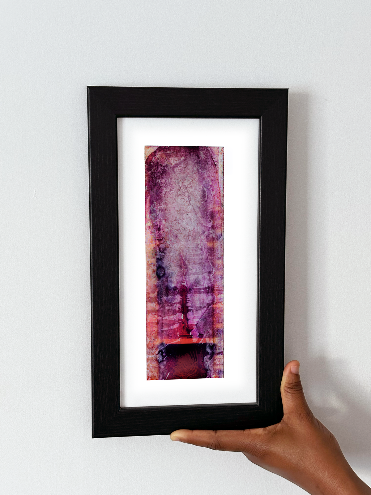 A hand holding a black-framed abstract art print with a white mat, featuring Naturalia II #015 against a plain white wall.

