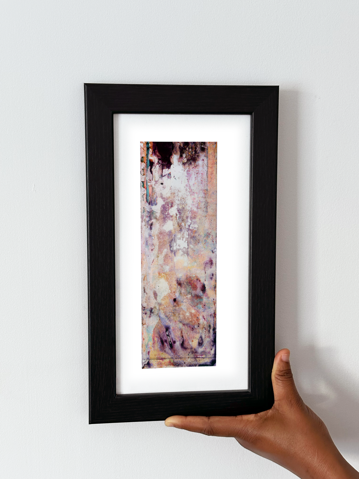 A hand holding a black-framed abstract art print with a white mat, featuring Naturalia II #020 against a plain white wall.

