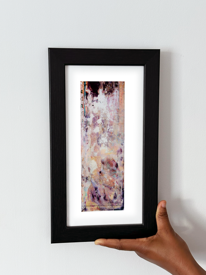 A hand holding a black-framed abstract art print with a white mat, featuring Naturalia II #020 against a plain white wall.
