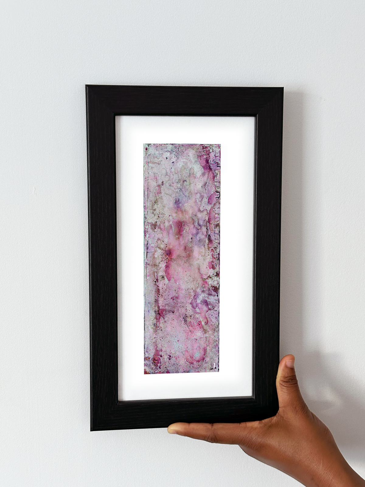 A hand holding a black-framed abstract art print with a white mat, featuring Naturalia II #021 against a plain white wall.
