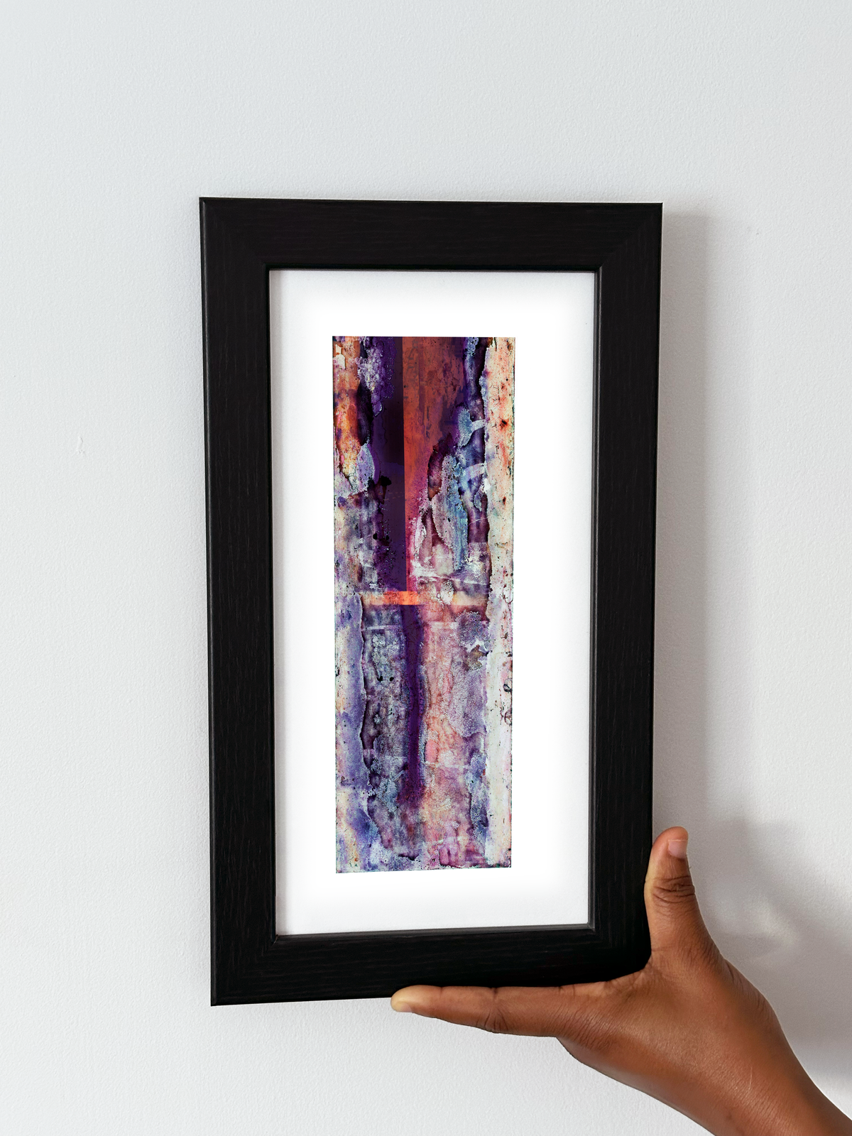 A hand holding a black-framed abstract art print with a white mat, featuring Naturalia II #024 against a plain white wall.

