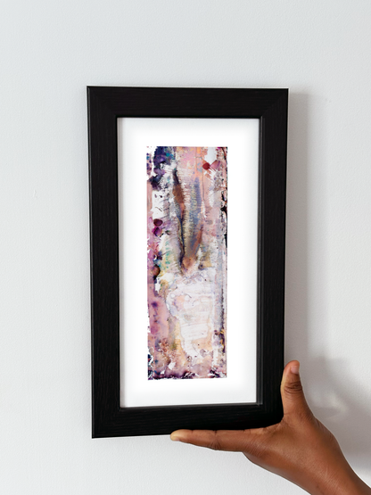 A hand holding a black-framed abstract art print with a white mat, featuring Naturalia II #027 against a plain white wall.
