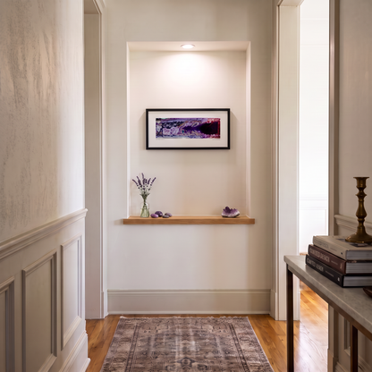 A framed horizontal art print with purple and black tones is displayed on a white wall in a hallway niche above a wooden shelf with decorative items. The scene includes a patterned rug on hardwood flooring, with a console table visible on the right.