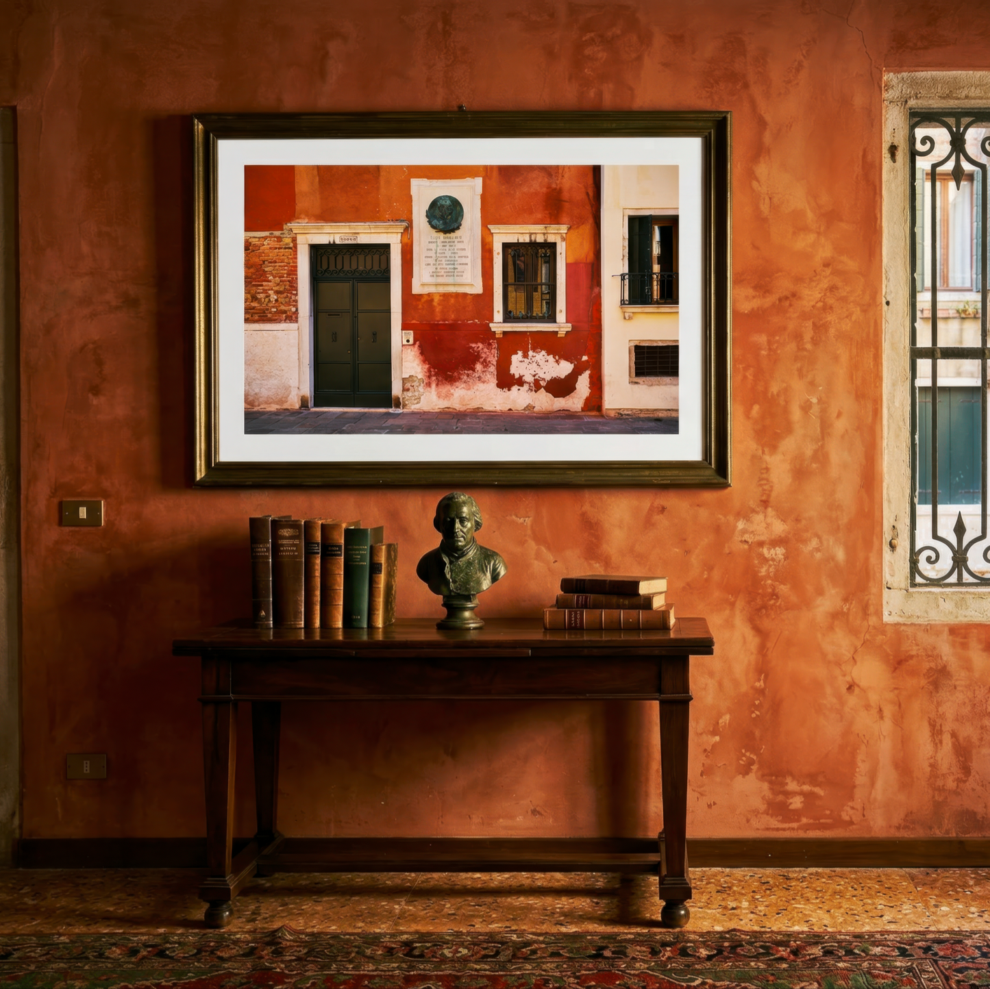 Room with orange walls, a framed photograph of a Venice doorway, a table with books and a bust, and a window with decorative iron bars.