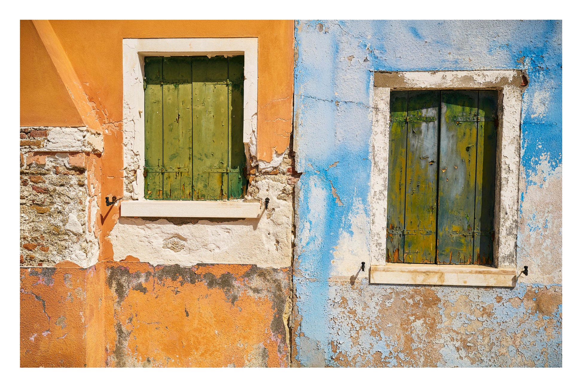 Two windows with green shutters on a wall with peeling paint in two different colors.