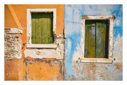 Two windows with green shutters on a wall with peeling paint in two different colors.