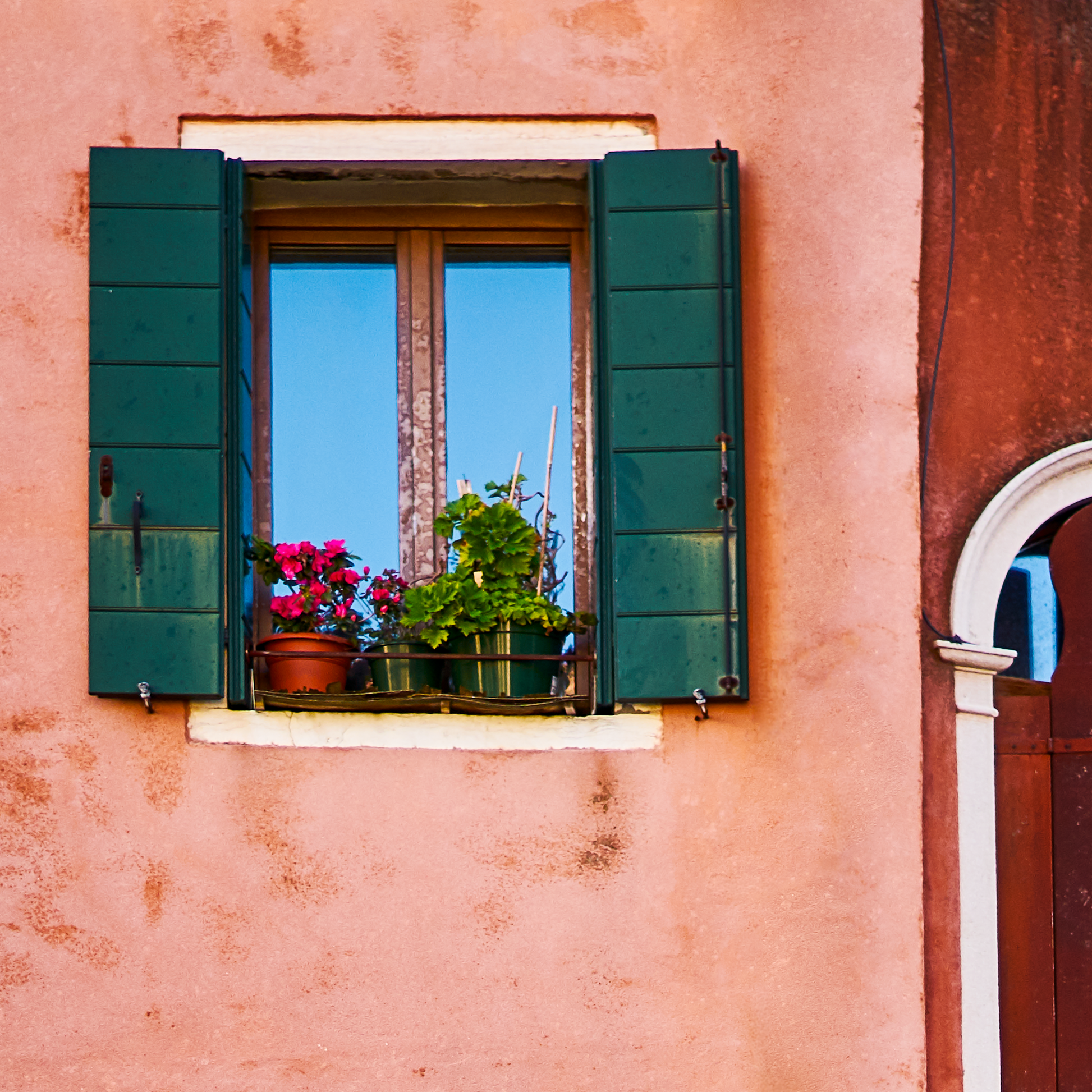 Window with green shutters on a pink wall, featuring potted plants.