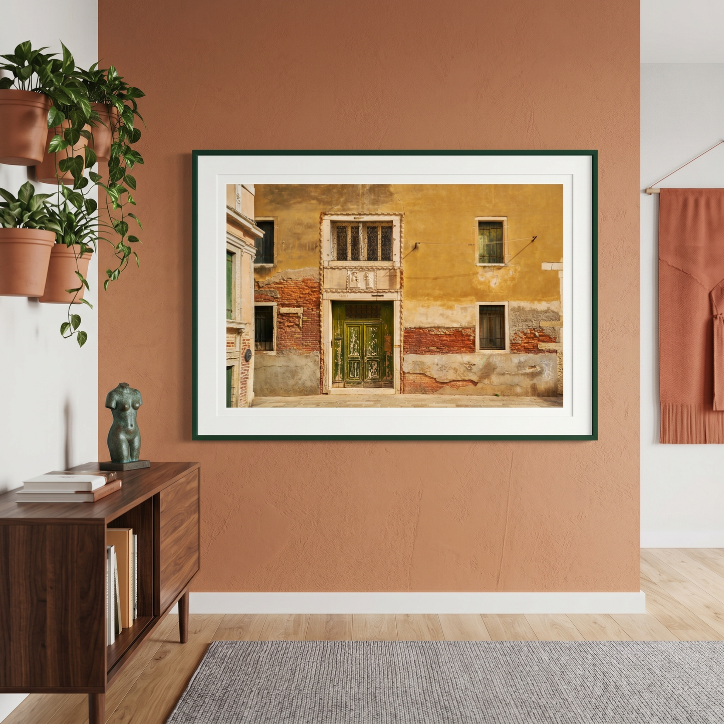 Framed artwork of a colorful building on a wall in a room with a wooden side table and plants.